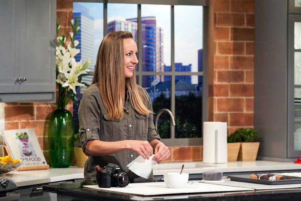 Lindsay Ostrom standing in a TV kitchen with a camera on the counter. 