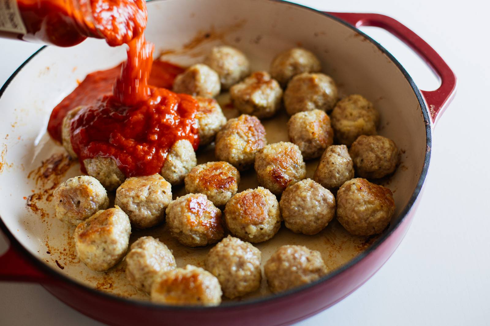 Pouring red sauce on meatballs in a skillet.