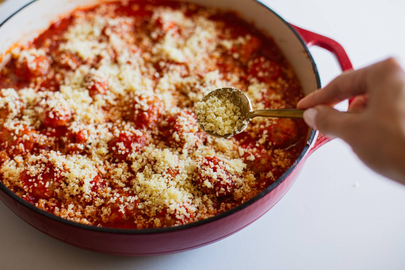 Sprinkling breadcrumbs on a skillet of meatballs in red sauce.