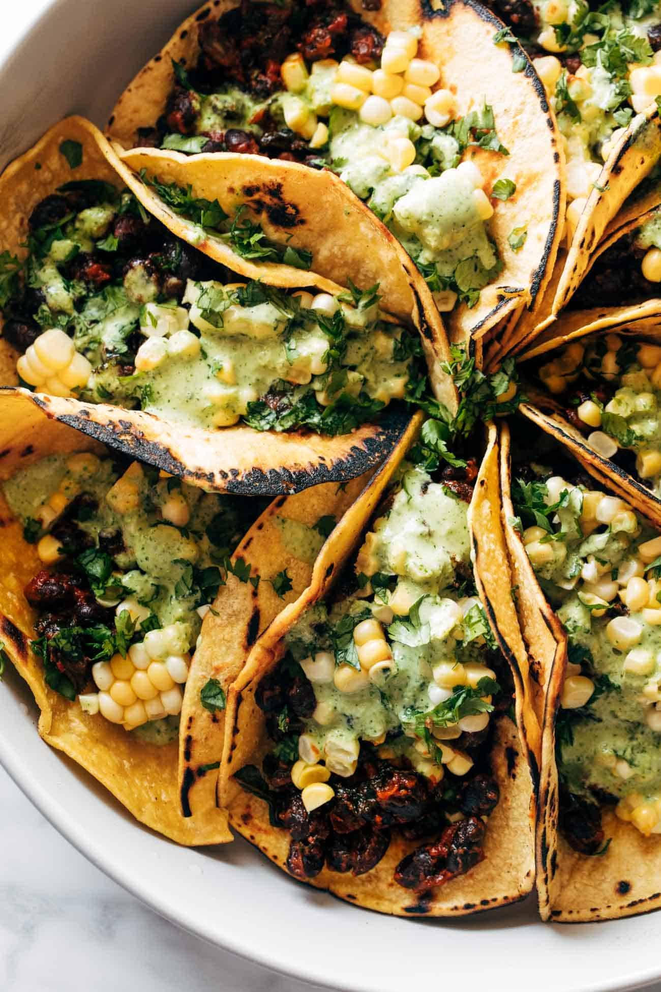 Smoky beans and greens tacos tucked together in a pan and topped with corn and Aji Verde sauce. 