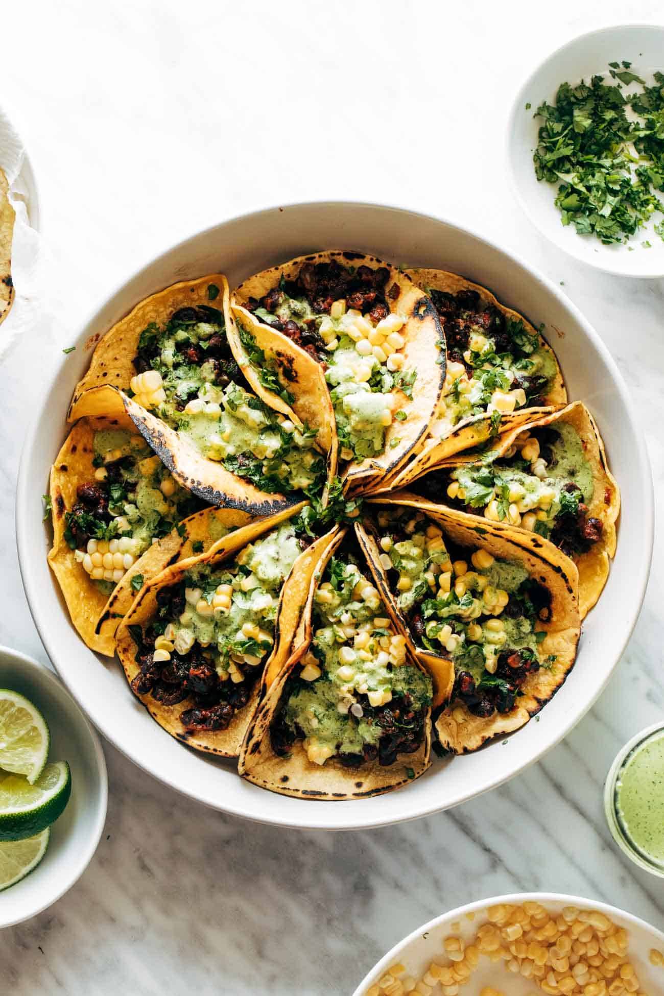 Smoky beans and greens tacos in a circular pattern on a plate and topped with Aji Verde sauce and corn. 