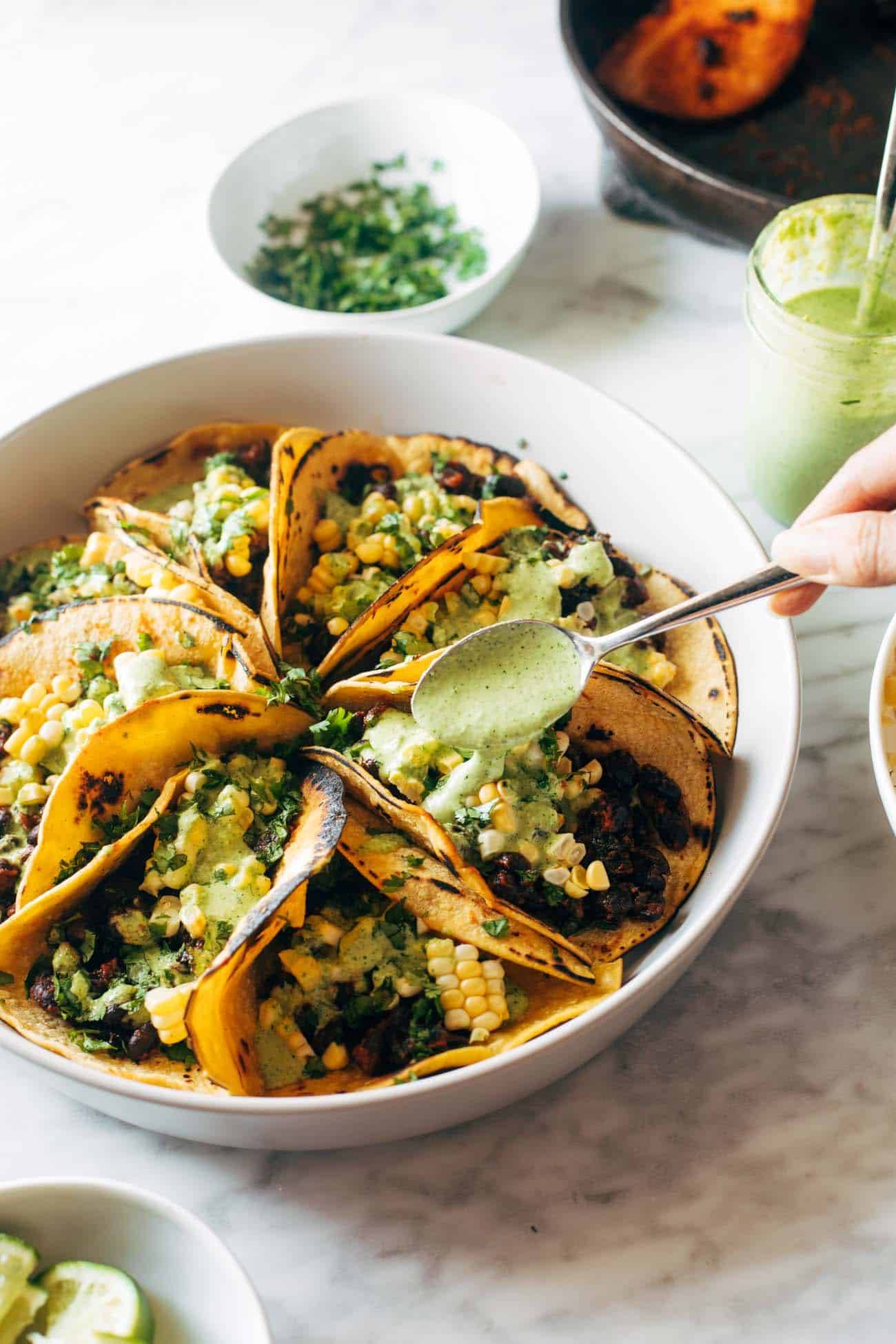 Smoky beans and greens tacos on a plate with corn and aji verde sauce being drizzled on top with a metal spoon. 