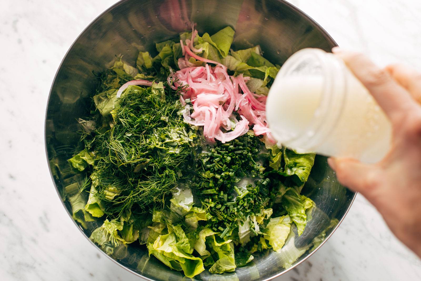 Pouring salad dressing on a bowl of greens and herbs.