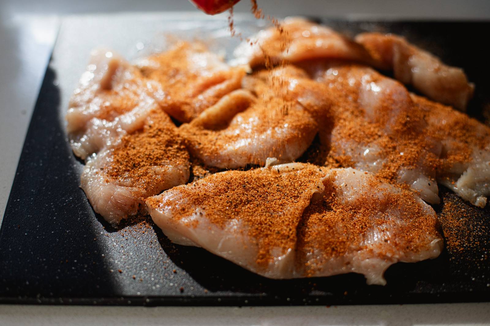 Sprinkling spice mix on raw chicken on a cutting board.