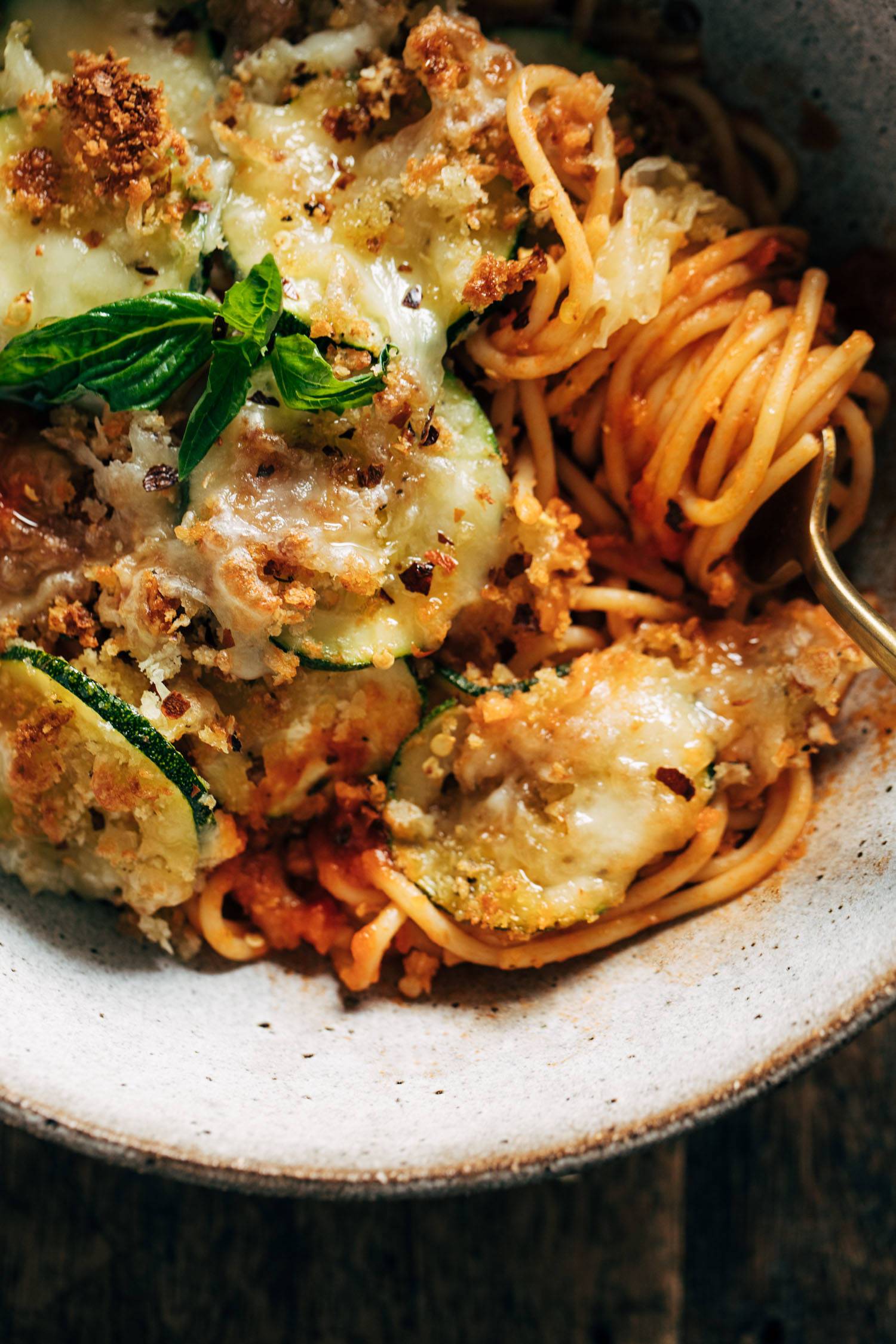 Close-up of spaghetti with crispy zucchini in a bowl with melted cheese and fresh basil on top. A fork is in the bowl twirling the spaghetti. 