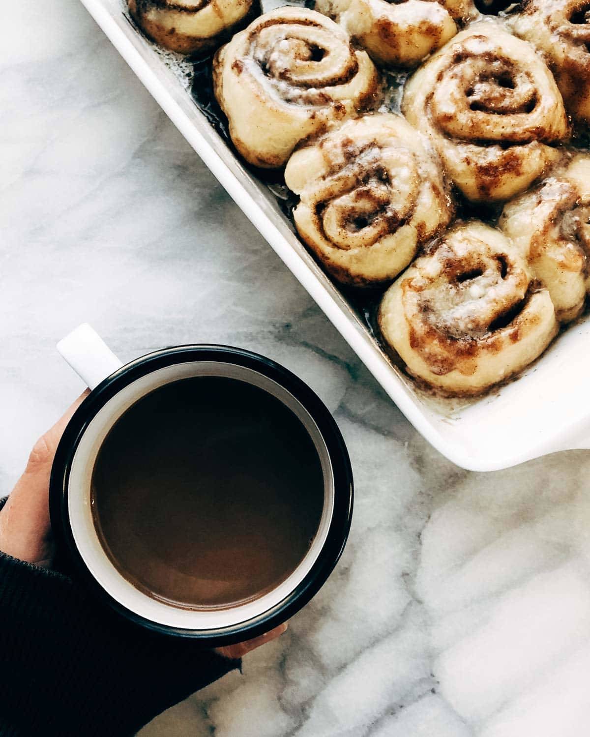 A mug of coffee alongside a pan of cinnamon rolls.