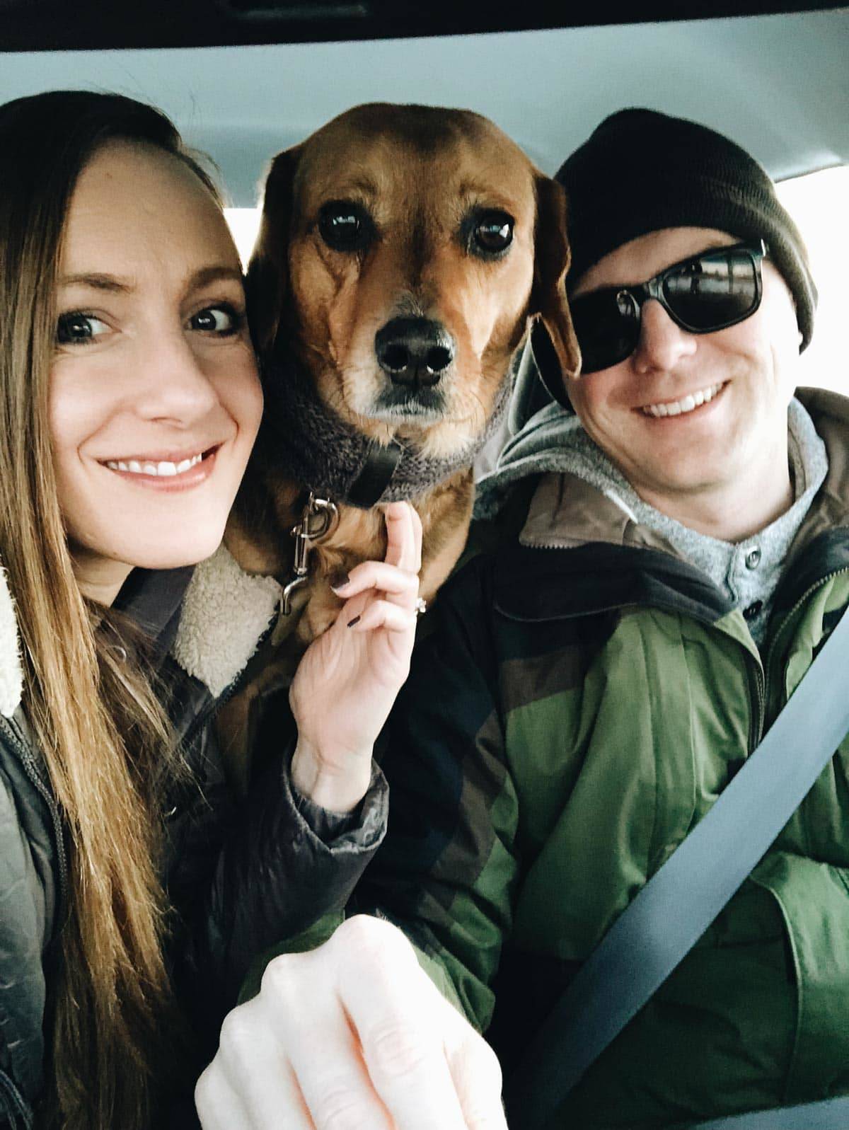 A smiling man and woman are in the front seat of a vehicle with a dog between them.