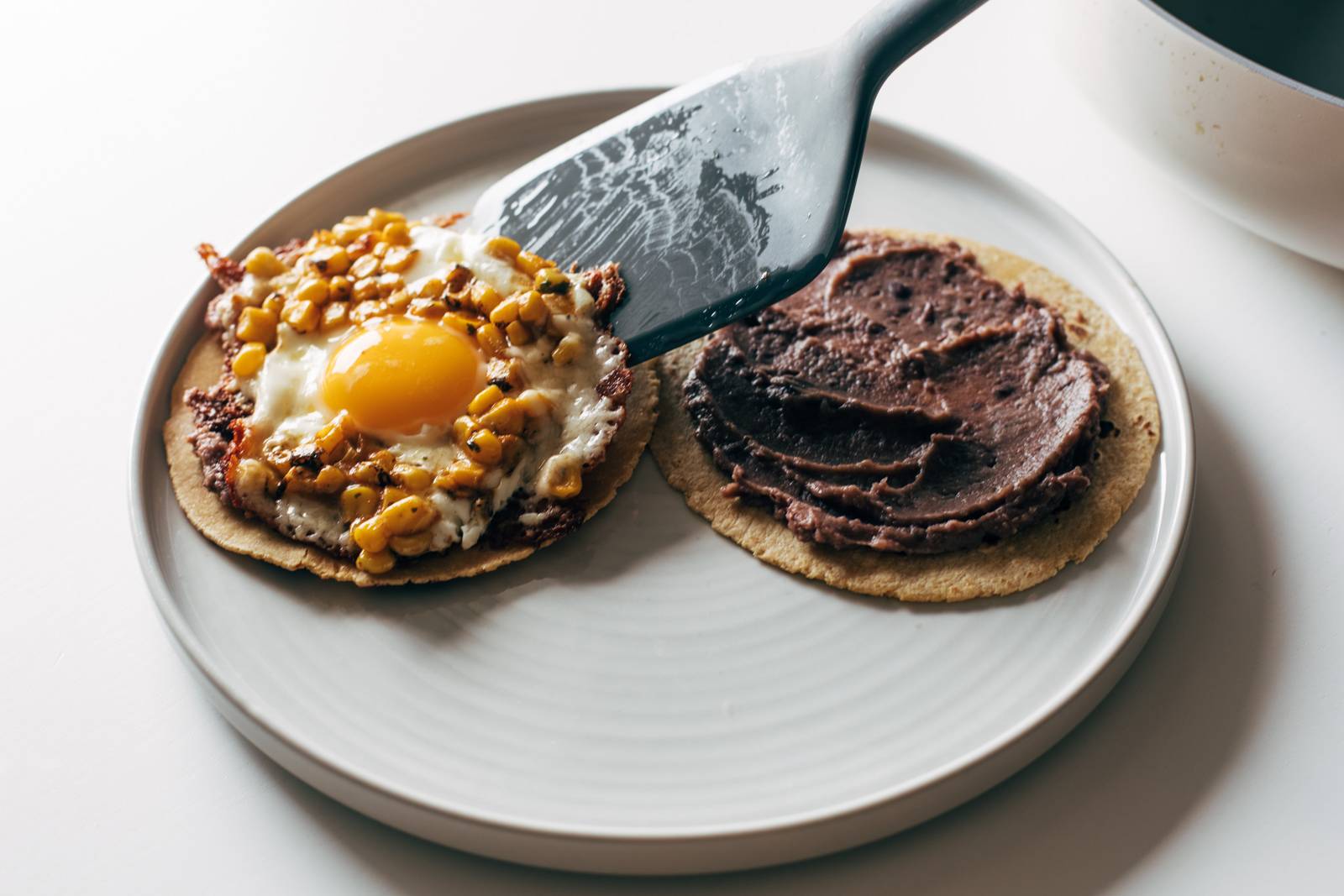 Placing fried egg and corn on a tortilla with refried beans spread on top.