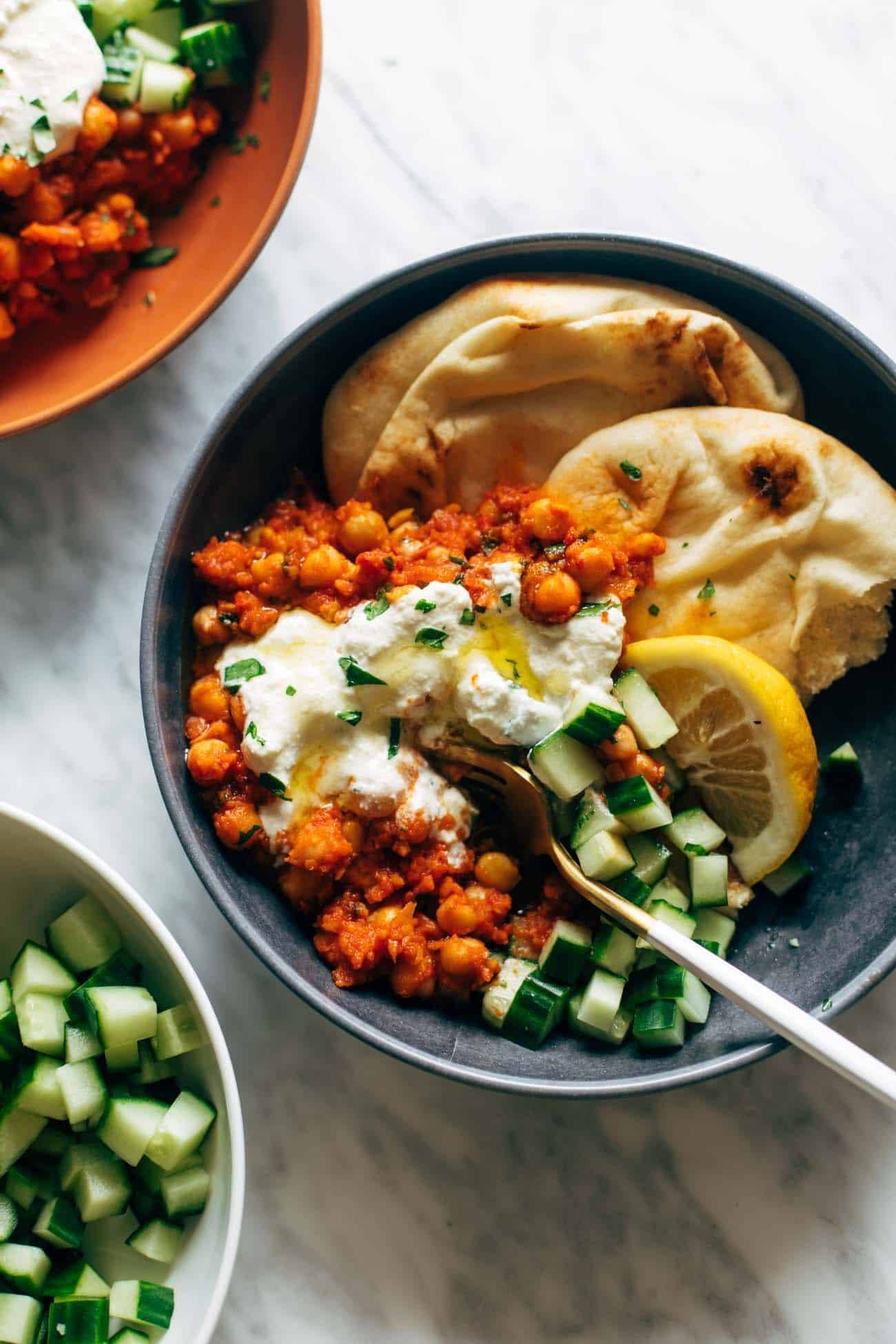 Harissa chickpeas with whipped feta in a bowl with a fork, cucumbers, lemon, and naan. 