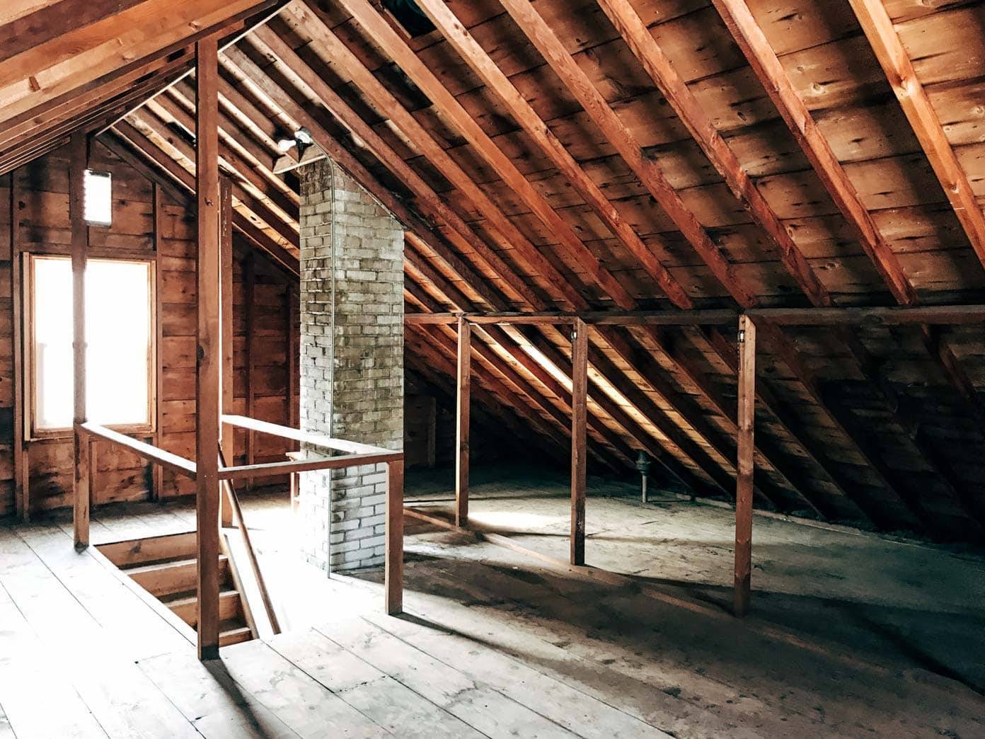 A well lit attic has a window and a chimney in it.