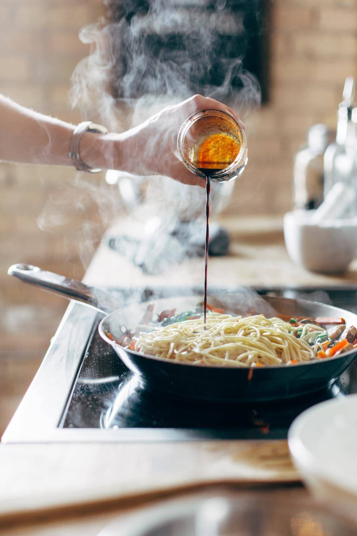 Pouring sauce into a pan with noodles and veggies.