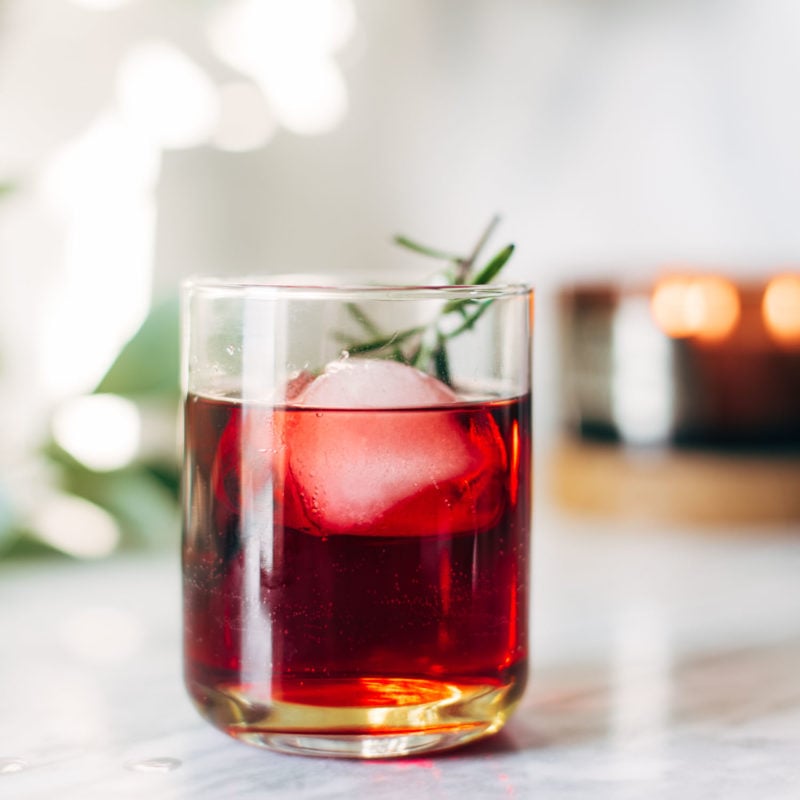 A clear cocktail glass with red liquid, a large ice cube, and a sprig of rosemary.