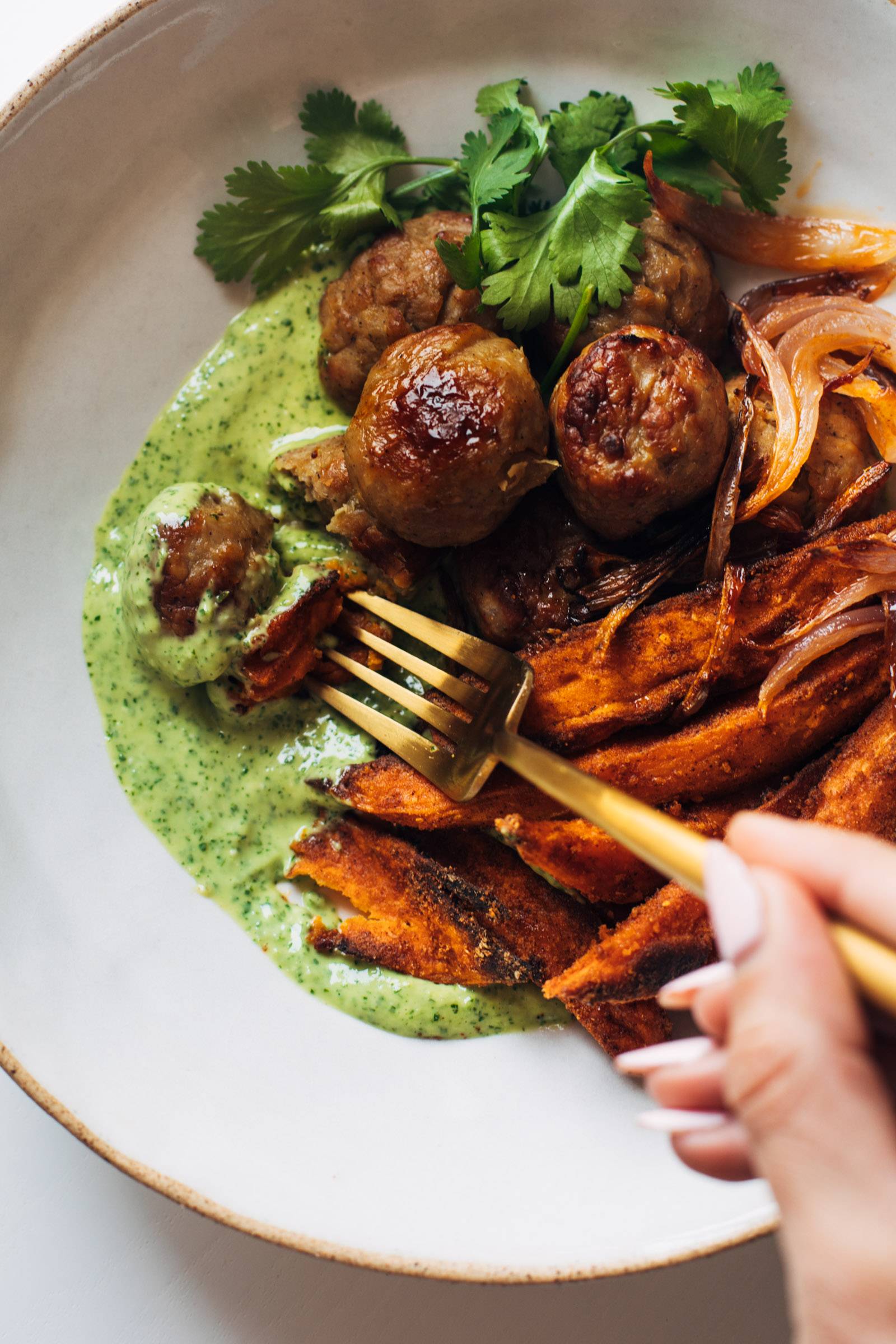 White hand holding a gold fork and eating a meatball in mojo bowls.