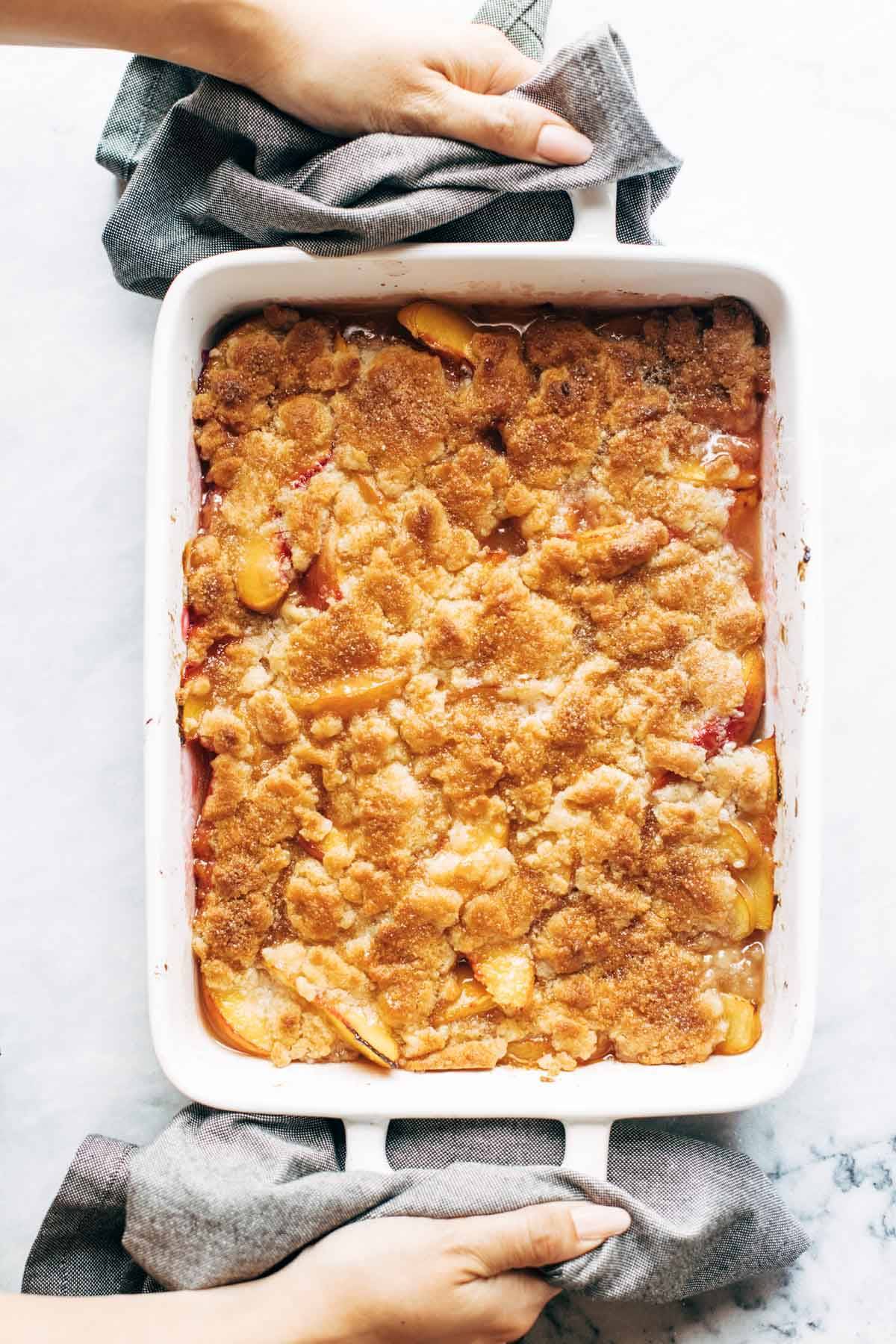 Cooked peach cobbler in a dish. White hands are holding the dish with a blue napkin. 