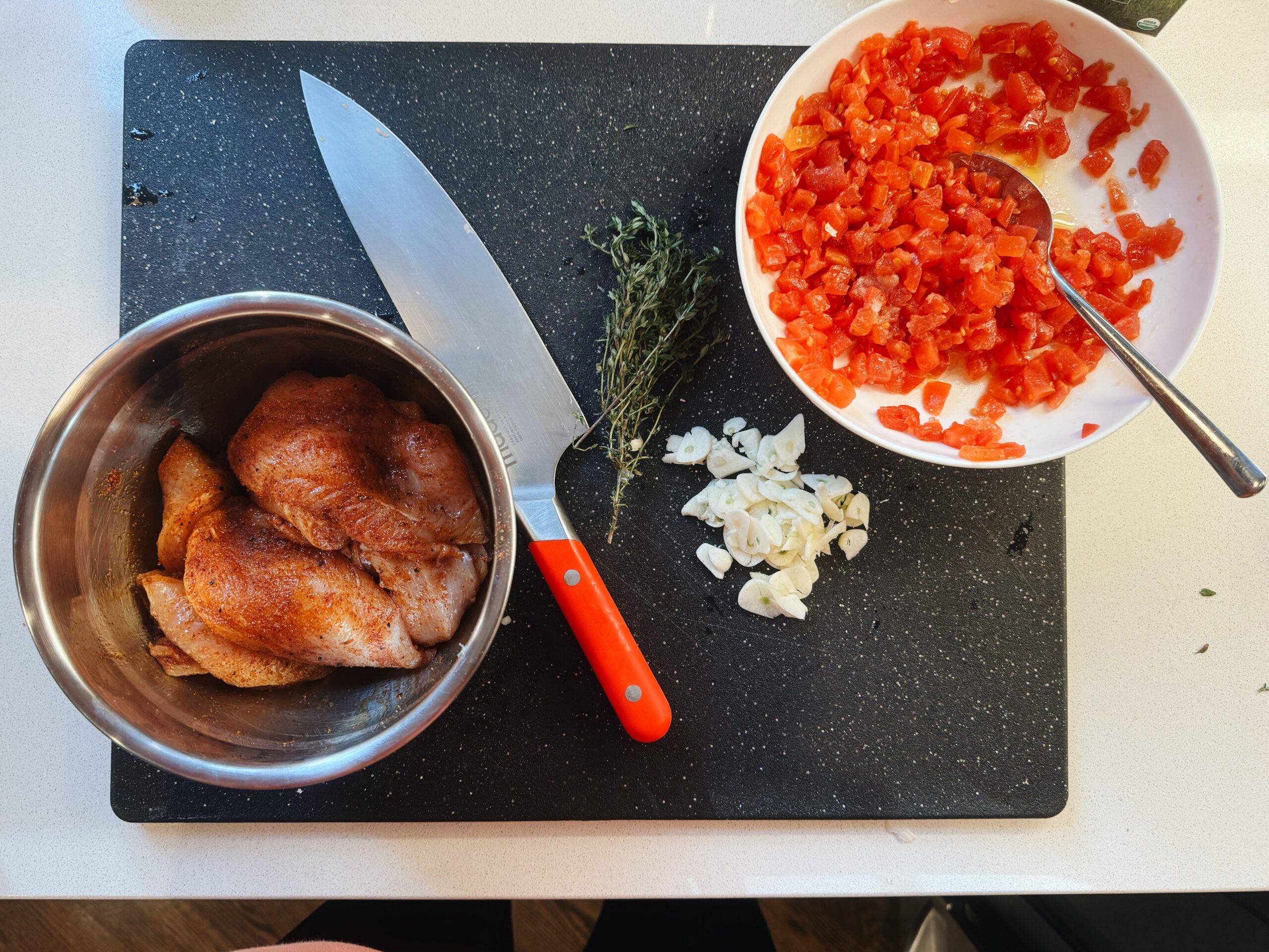 Raw chicken in a bowl next to a bowl of tomatoes and chopped garlic and herbs,