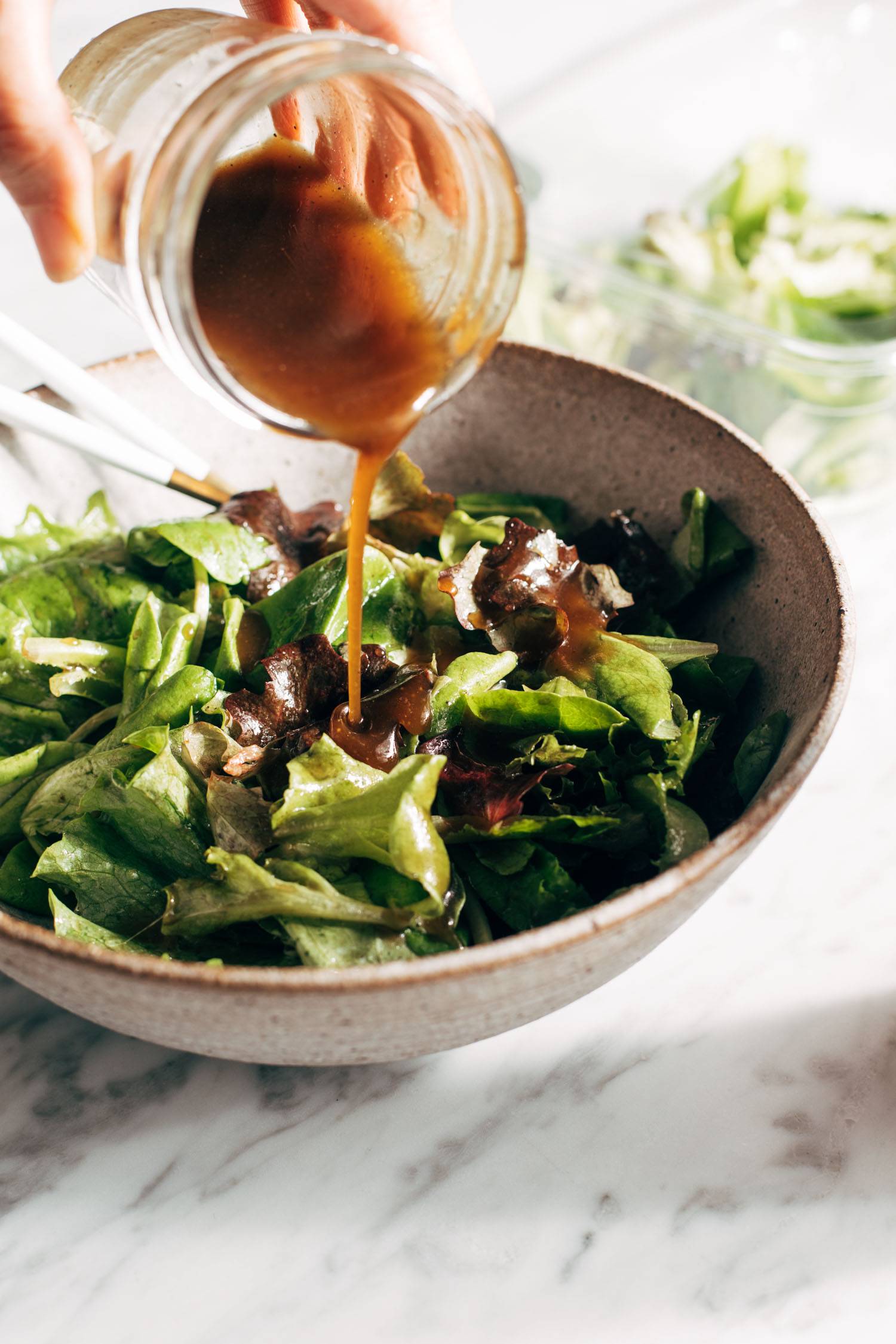 White hand pouring dressing over a bowl of salad greens.