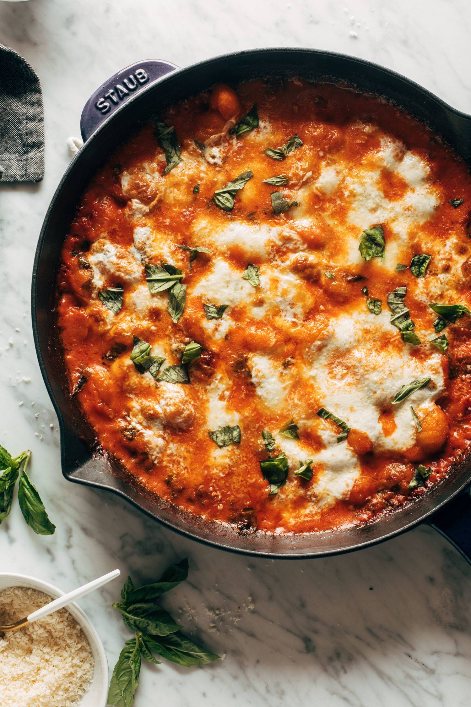 Three cheese baked gnocchi sprinkled with fresh basil on top. The gnocchi is in a cast iron pan and there's fresh basil and grated parmesan cheese in a bowl on the side. 