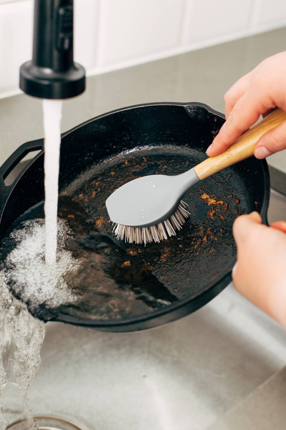 Scrubbing crusted cast iron skillet under water with a brush.