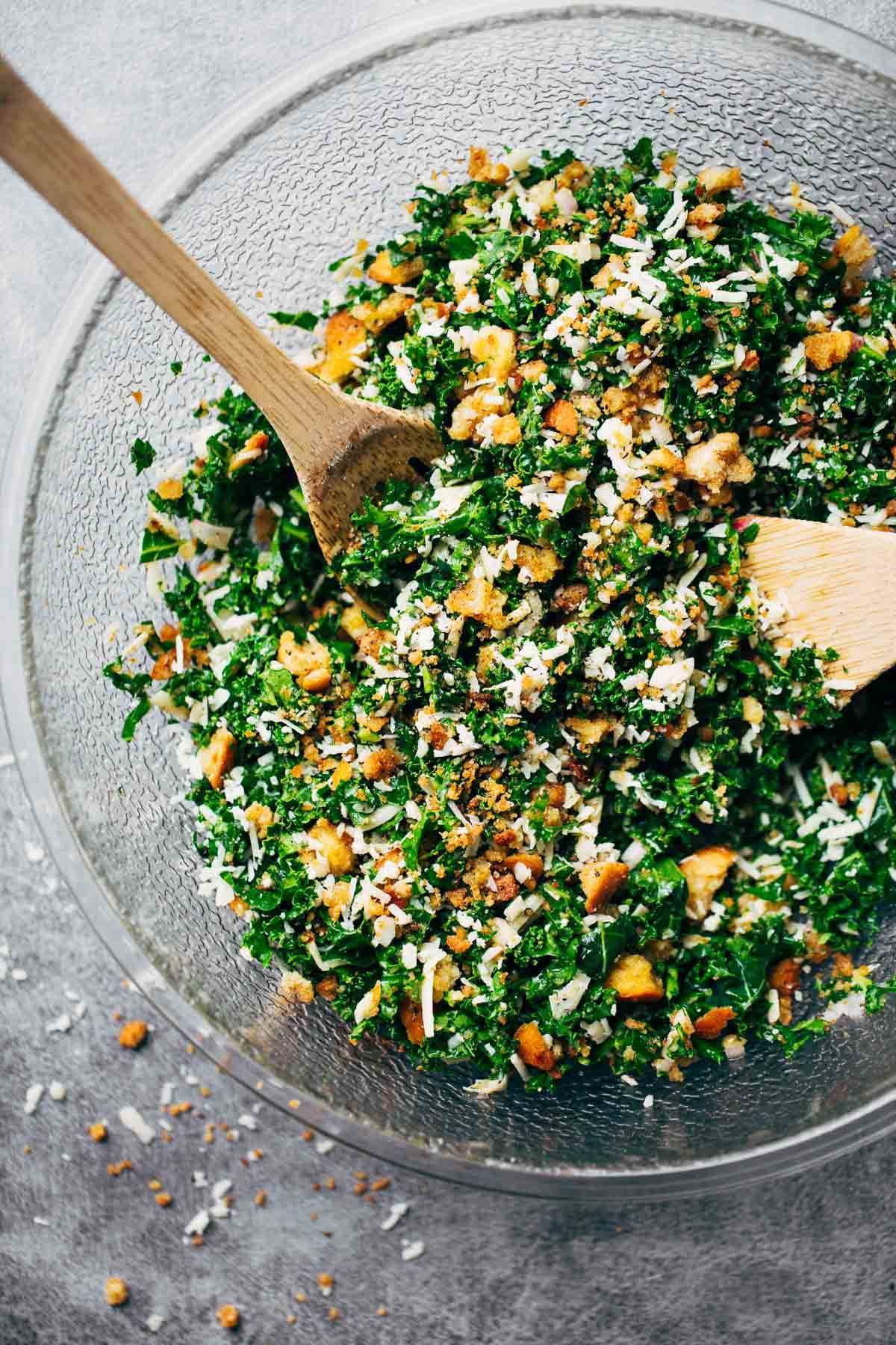 Kale Salad with Parmesan cheese, homemade breadcrumbs, and a lemon olive oil dressing in a mixing bowl with spoons.